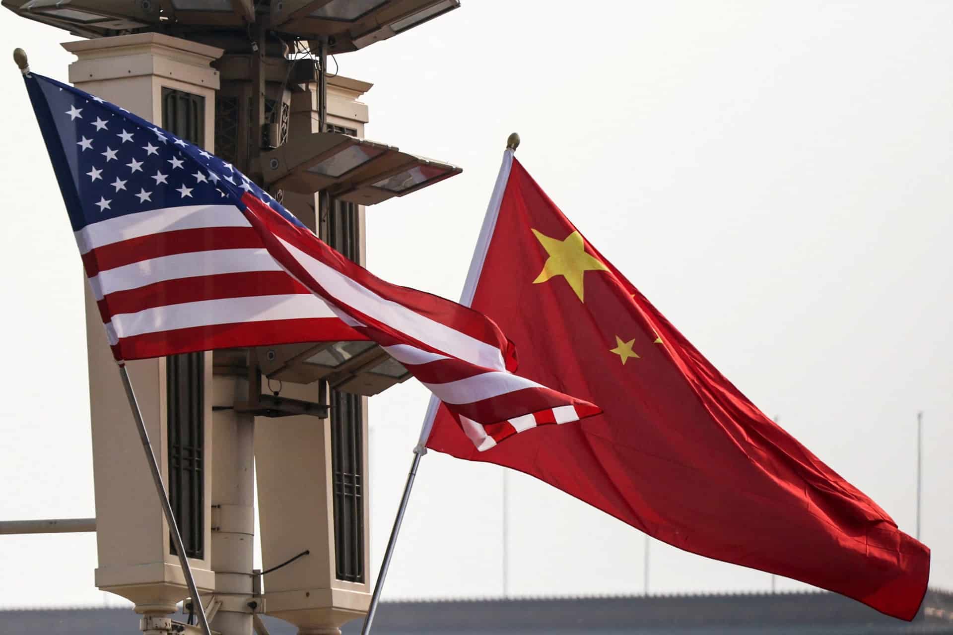 The American flag and the Chinese flag wave side by side in front of a beige guard tower or surveillance structure, highlighting tensions over memory chips and China memory firms against a pale sky.