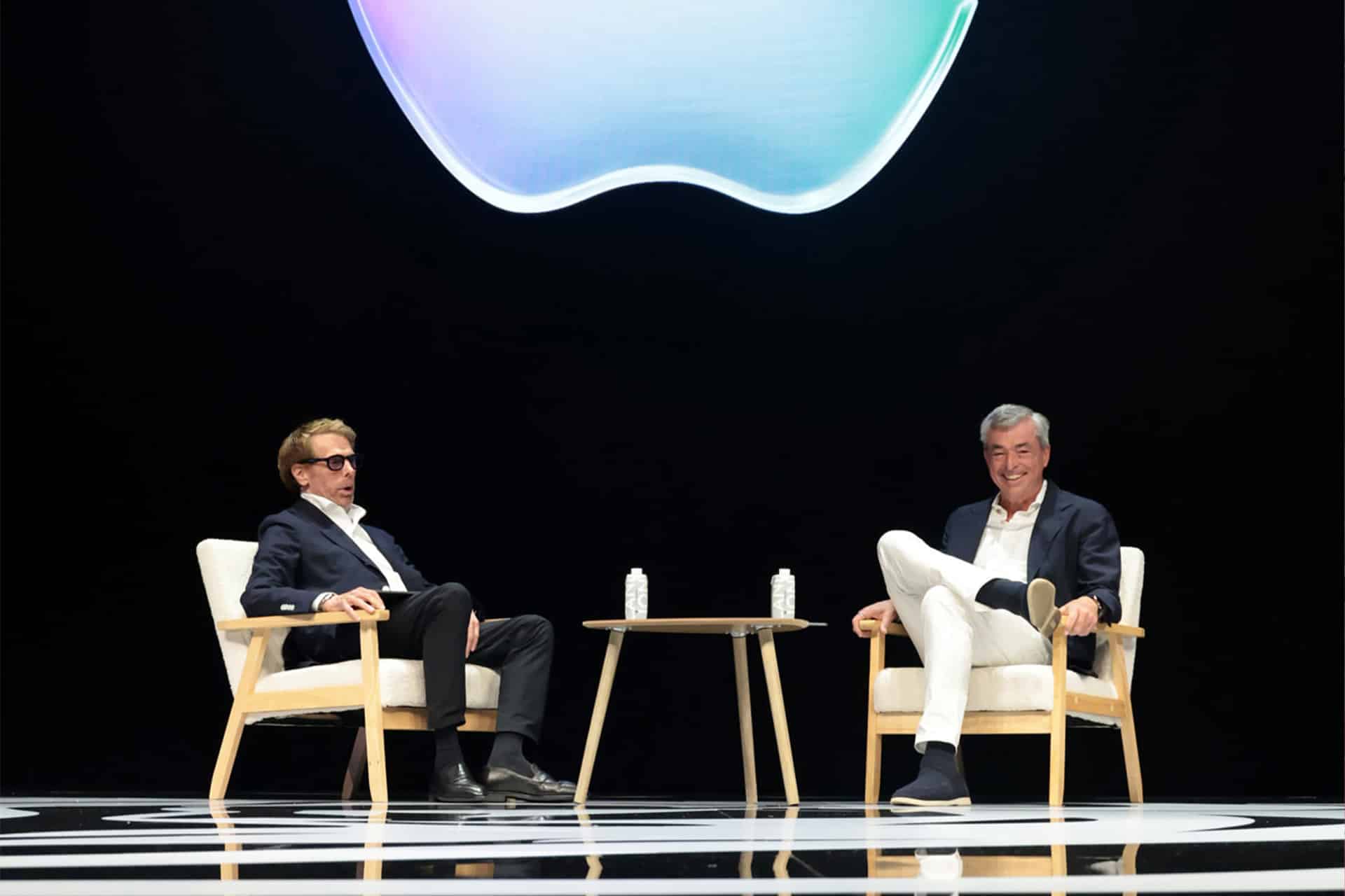 Two men sit and talk on stage in armchairs, a small table between them, under a large Apple logo projected above, discussing Apple Services at Cannes Lions. Both are smiling in suits against a black background.