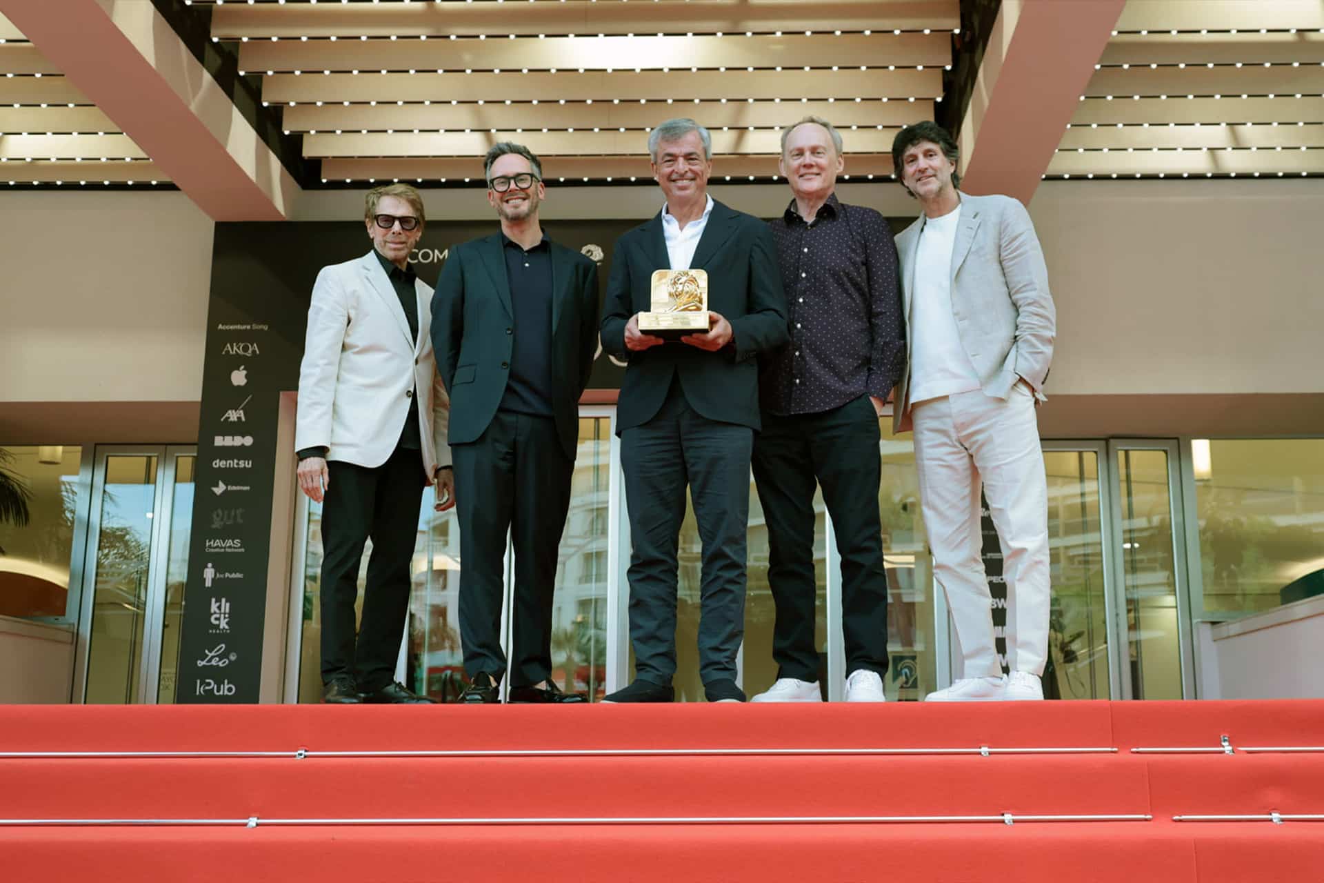 Five men stand together on a red carpeted staircase, dressed in suits and casual attire. The man in the center holds a golden award, with Eddy Cue from Apple Services smiling among them, and a modern building entrance in the background.