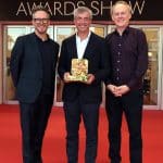 Three men stand smiling on a red carpet at Cannes Lions in front of glass doors. The man in the center, Eddy Cue of Apple Services, holds a golden award. Event sponsor logos fill the background. The atmosphere is celebratory and formal.