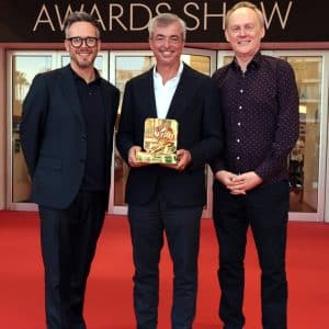 Three men stand smiling on a red carpet at Cannes Lions in front of glass doors. The man in the center, Eddy Cue of Apple Services, holds a golden award. Event sponsor logos fill the background. The atmosphere is celebratory and formal.