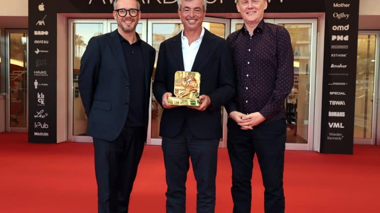 Three men stand smiling on a red carpet at Cannes Lions in front of glass doors. The man in the center, Eddy Cue of Apple Services, holds a golden award. Event sponsor logos fill the background. The atmosphere is celebratory and formal.