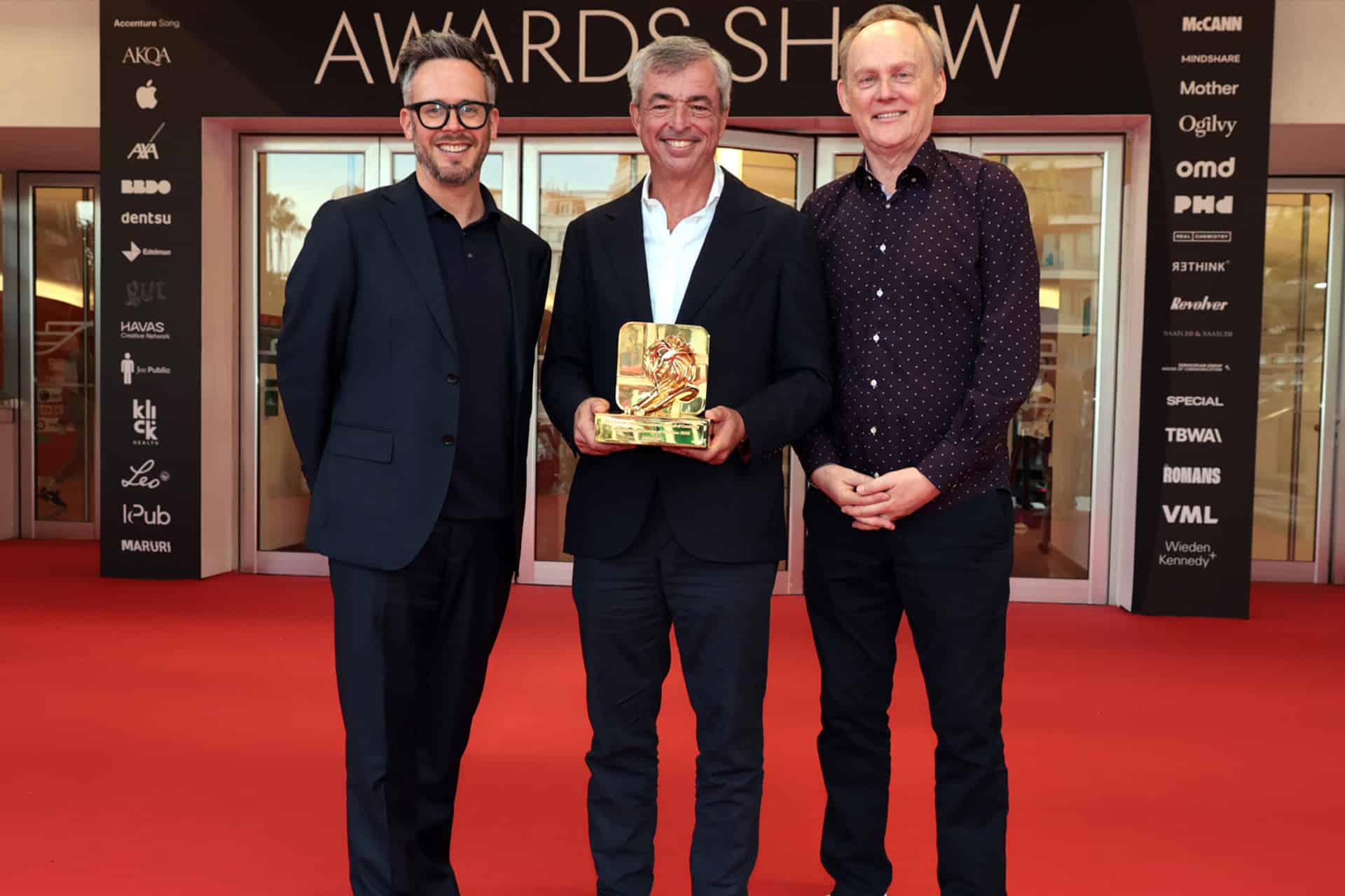 Three men stand smiling on a red carpet at Cannes Lions in front of glass doors. The man in the center, Eddy Cue of Apple Services, holds a golden award. Event sponsor logos fill the background. The atmosphere is celebratory and formal.