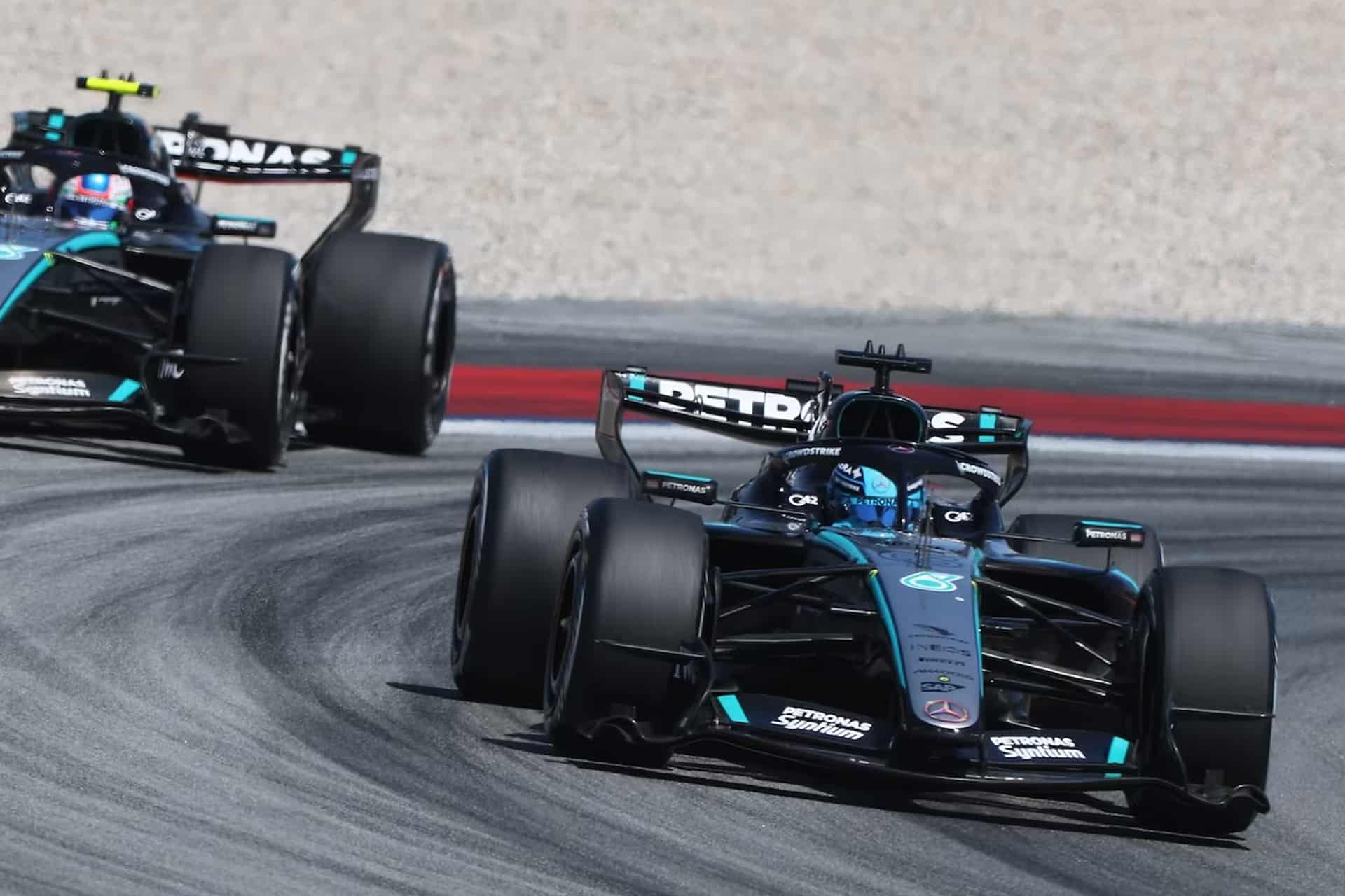 Two black Mercedes Formula 1 cars racing closely on a curved track at the Red Bull Ring, with the leading car taking the inside line while the second follows. Both display turquoise accents, Apple logos, and visible sponsor branding during the Austrian Grand Prix.