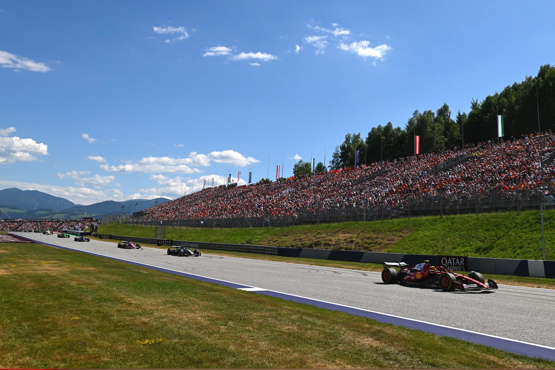 Formula 1 cars race on the iconic Red Bull Ring under a blue sky, with a large crowd of spectators filling the grandstands on a grassy hillside beside the circuit at the Austrian Grand Prix.