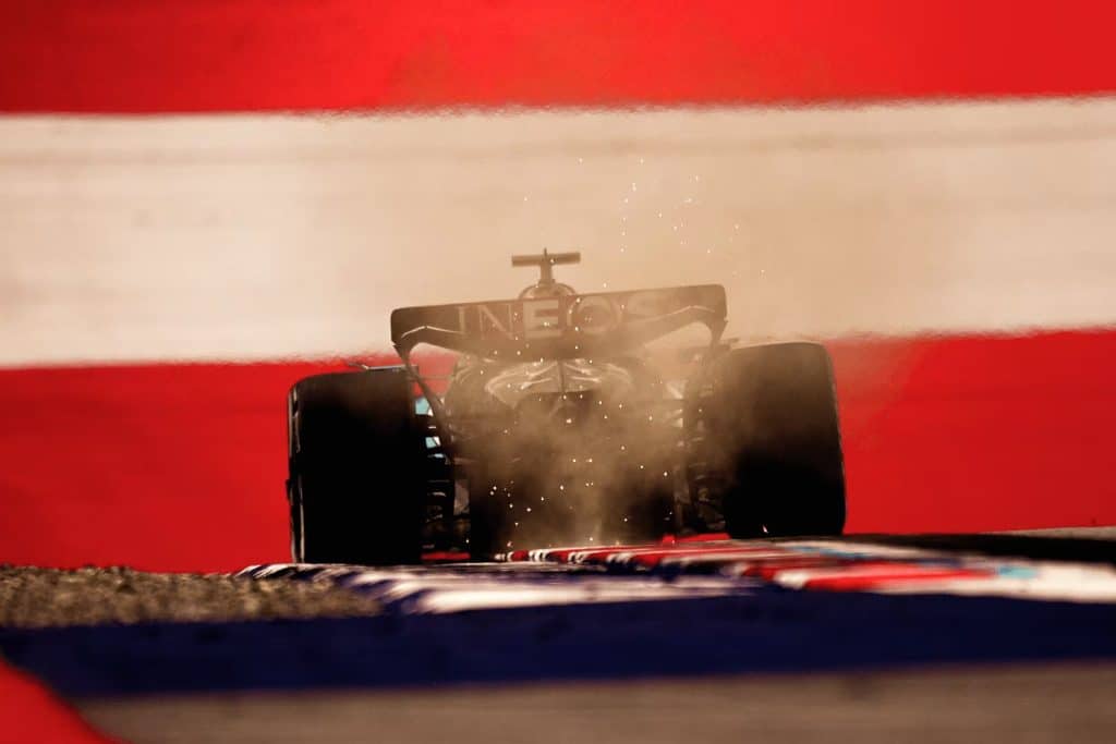 A Formula 1 car kicks up dust as it speeds along the track at the Austrian Grand Prix, backlit against a dramatic background of bold red and white stripes.