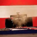 A Formula 1 car kicks up dust as it speeds along the track at the Austrian Grand Prix, backlit against a dramatic background of bold red and white stripes.