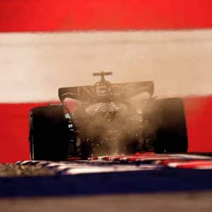 A Formula 1 car kicks up dust as it speeds along the track at the Austrian Grand Prix, backlit against a dramatic background of bold red and white stripes.