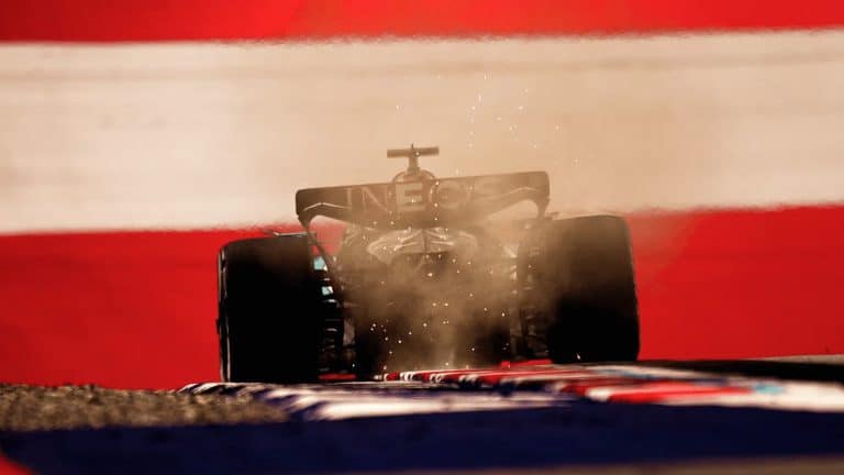 A Formula 1 car kicks up dust as it speeds along the track at the Austrian Grand Prix, backlit against a dramatic background of bold red and white stripes.