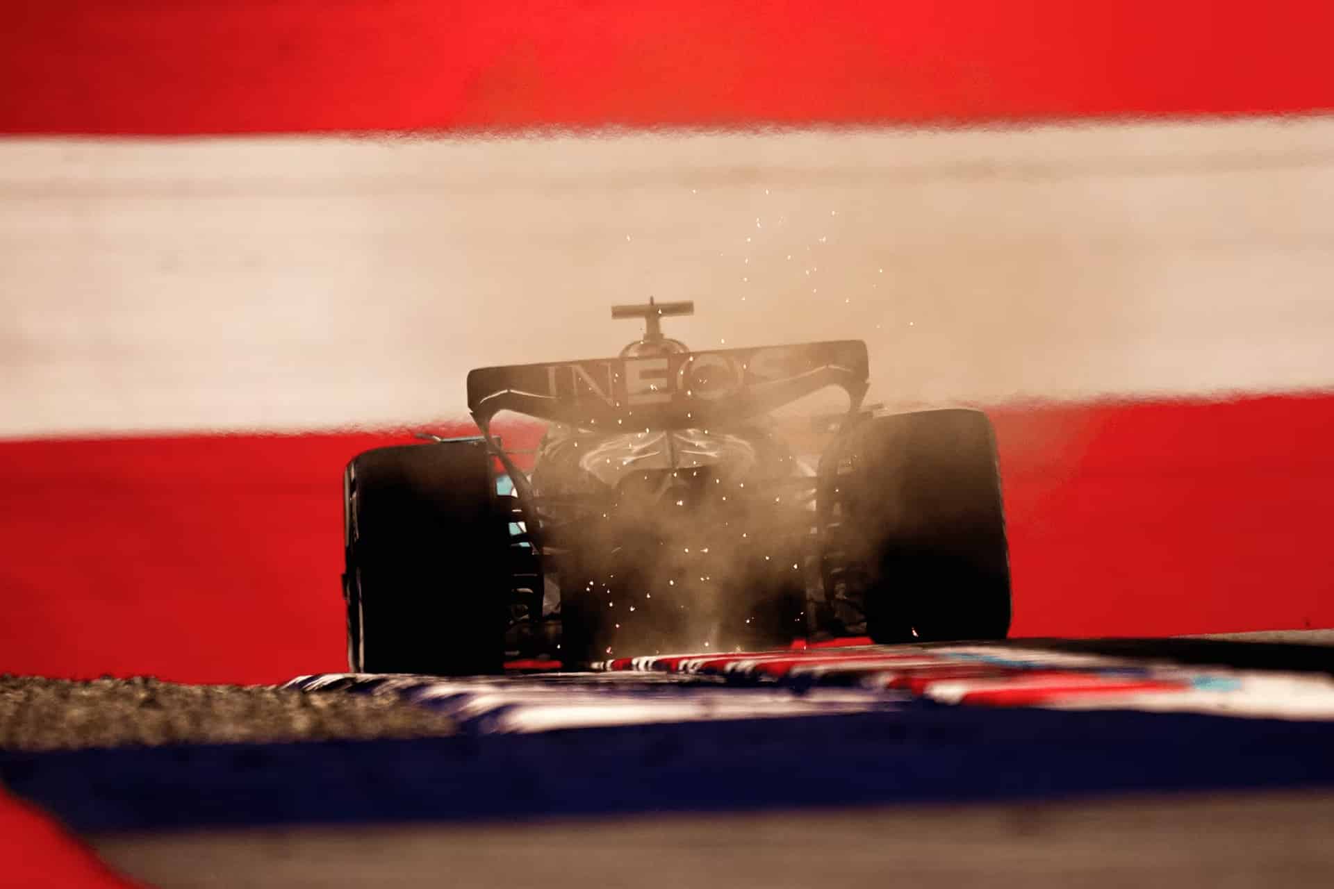 A Formula 1 car kicks up dust as it speeds along the track at the Austrian Grand Prix, backlit against a dramatic background of bold red and white stripes.