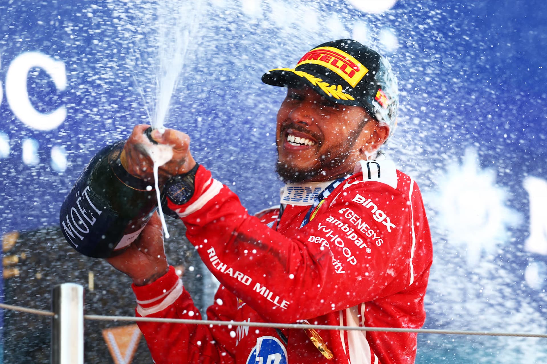 A racing driver in a red suit and cap sprays champagne and smiles in celebration on a podium at the Red Bull Ring, with droplets splashing everywhere against a blue background.