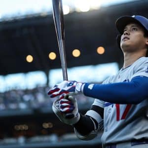 A baseball player in a Los Angeles uniform holds a bat and looks upward, preparing to bat during Friday Night Baseball. The stadium lights and crowd are visible in the background.