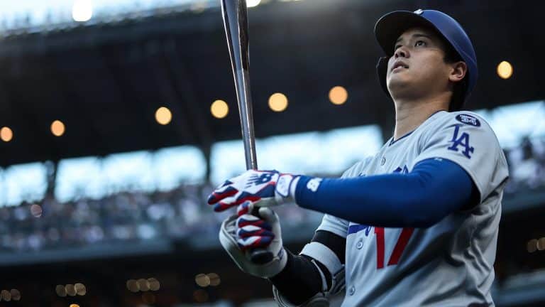 A baseball player in a Los Angeles uniform holds a bat and looks upward, preparing to bat during Friday Night Baseball. The stadium lights and crowd are visible in the background.