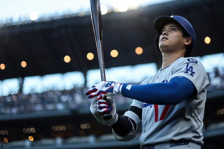 A baseball player in a Los Angeles uniform holds a bat and looks upward, preparing to bat during Friday Night Baseball. The stadium lights and crowd are visible in the background.