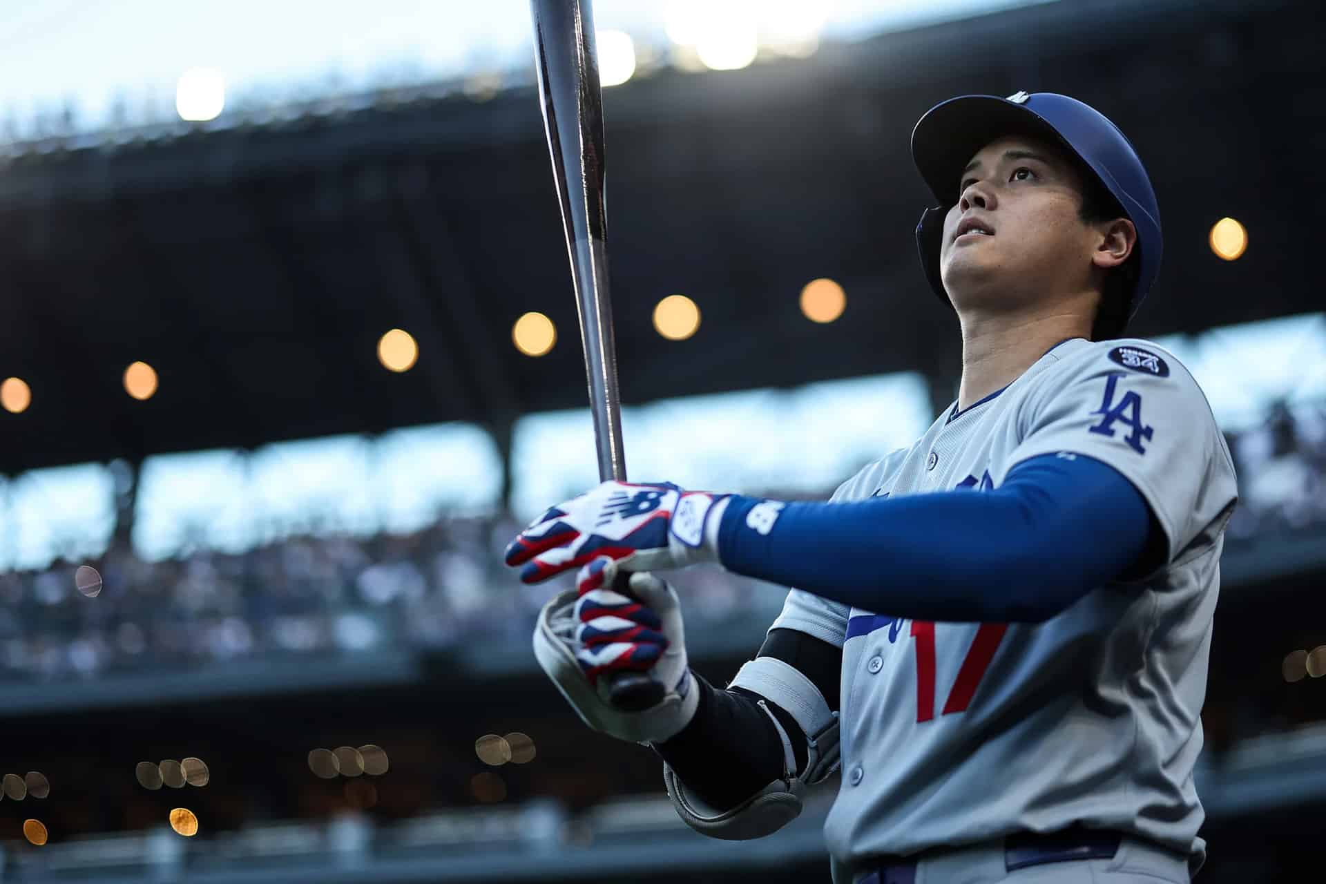 A baseball player in a Los Angeles uniform holds a bat and looks upward, preparing to bat during Friday Night Baseball. The stadium lights and crowd are visible in the background.