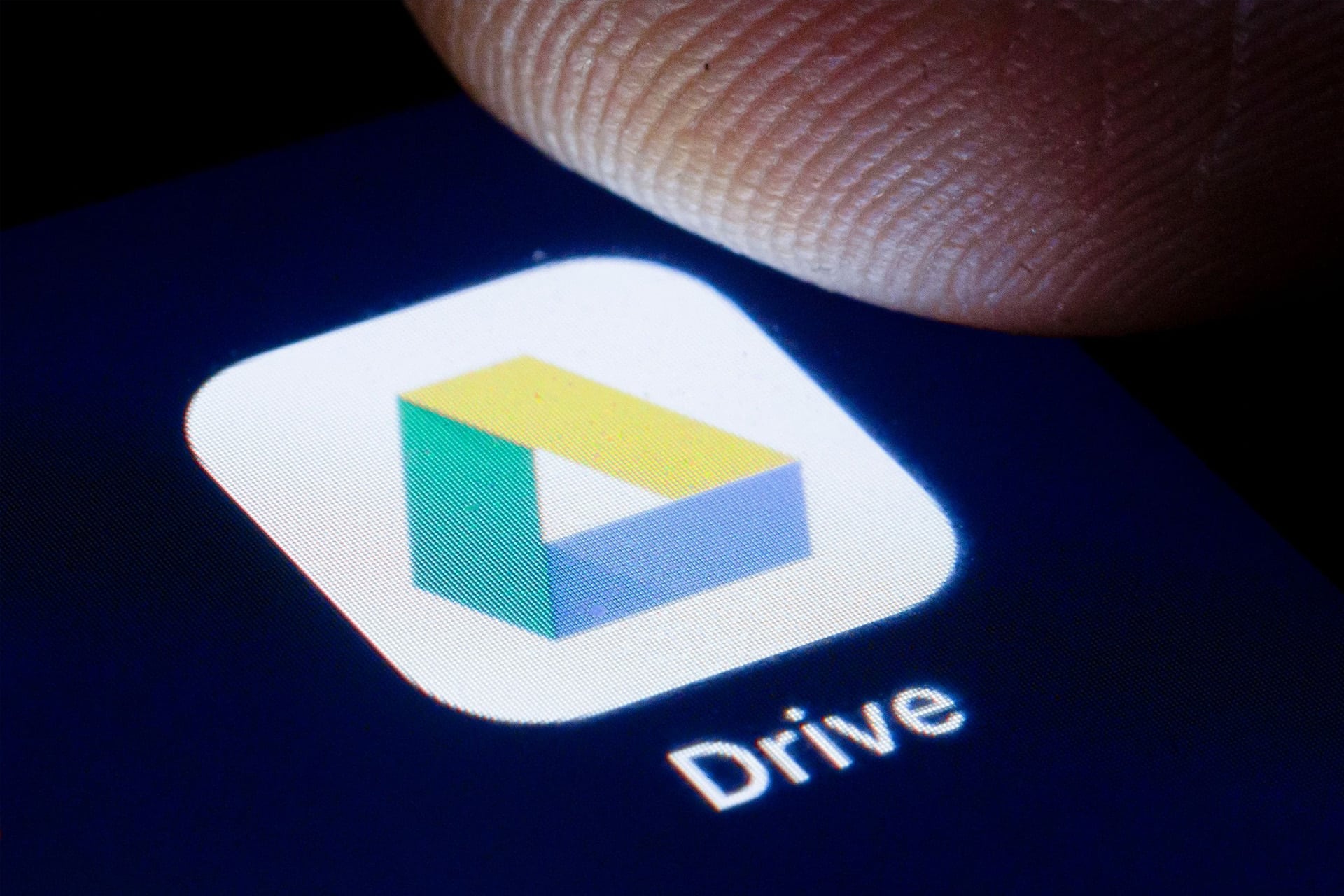 A close-up of a finger about to tap the Google Drive app icon on a screen, part of the Google Workspace suite that includes Gmail. The triangular icon in yellow, green, and blue has the word "Drive" below it on a Mac.