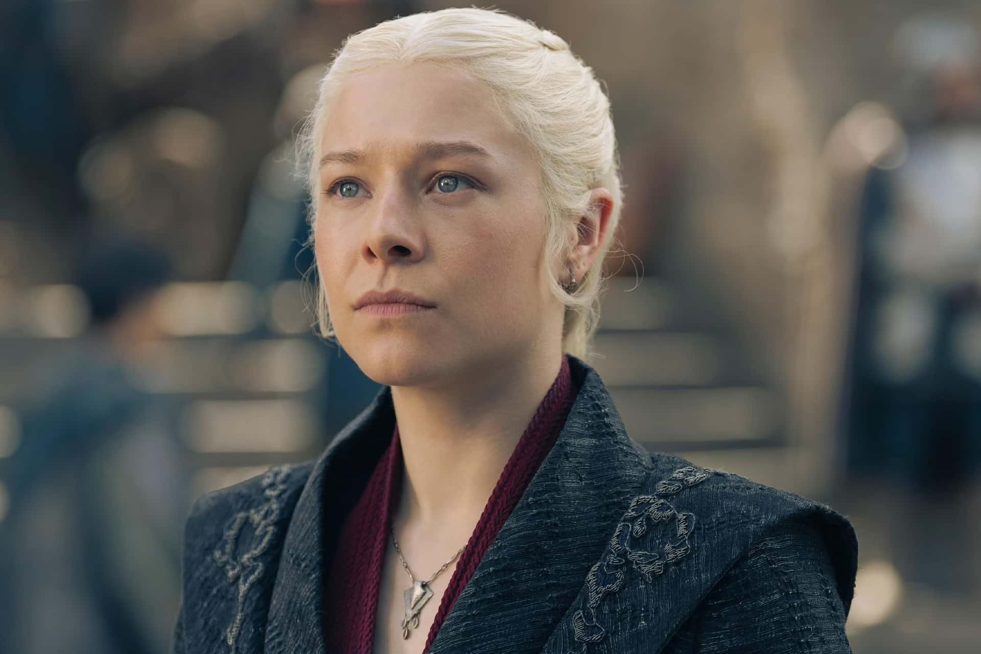 A person with pale blonde hair styled back, wearing a dark, embroidered jacket and a pendant necklace, stands outdoors with a serious expression—like a character from an Apple TV series. The background is blurred.