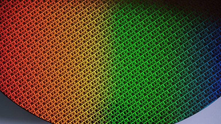 A close-up of a silicon wafer with a grid pattern reflecting a gradient of colors from orange to green to blue, highlighting the intricate design found in Apple chips and advanced M-series chips.