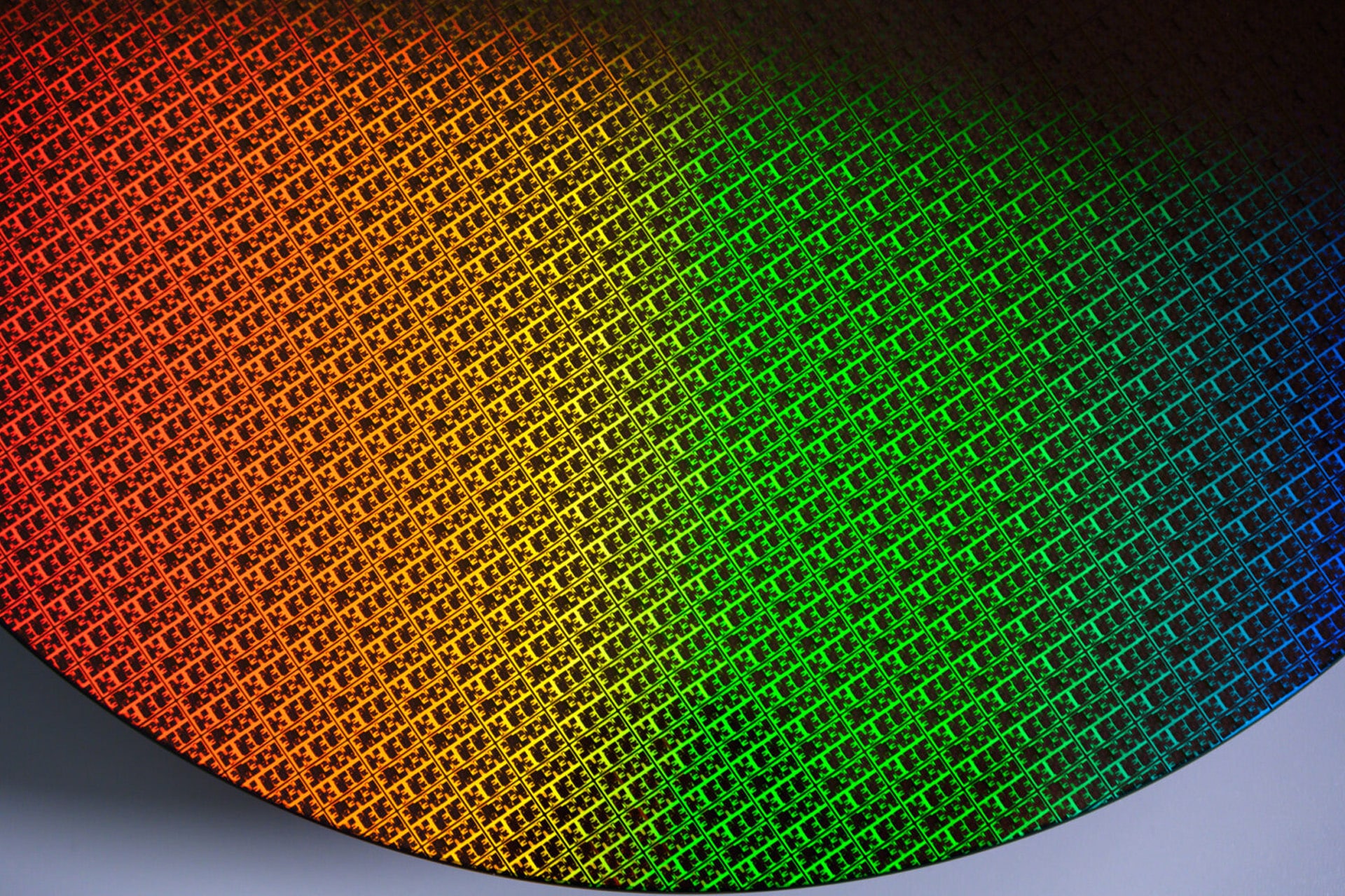 A close-up of a silicon wafer with a grid pattern reflecting a gradient of colors from orange to green to blue, highlighting the intricate design found in Apple chips and advanced M-series chips.