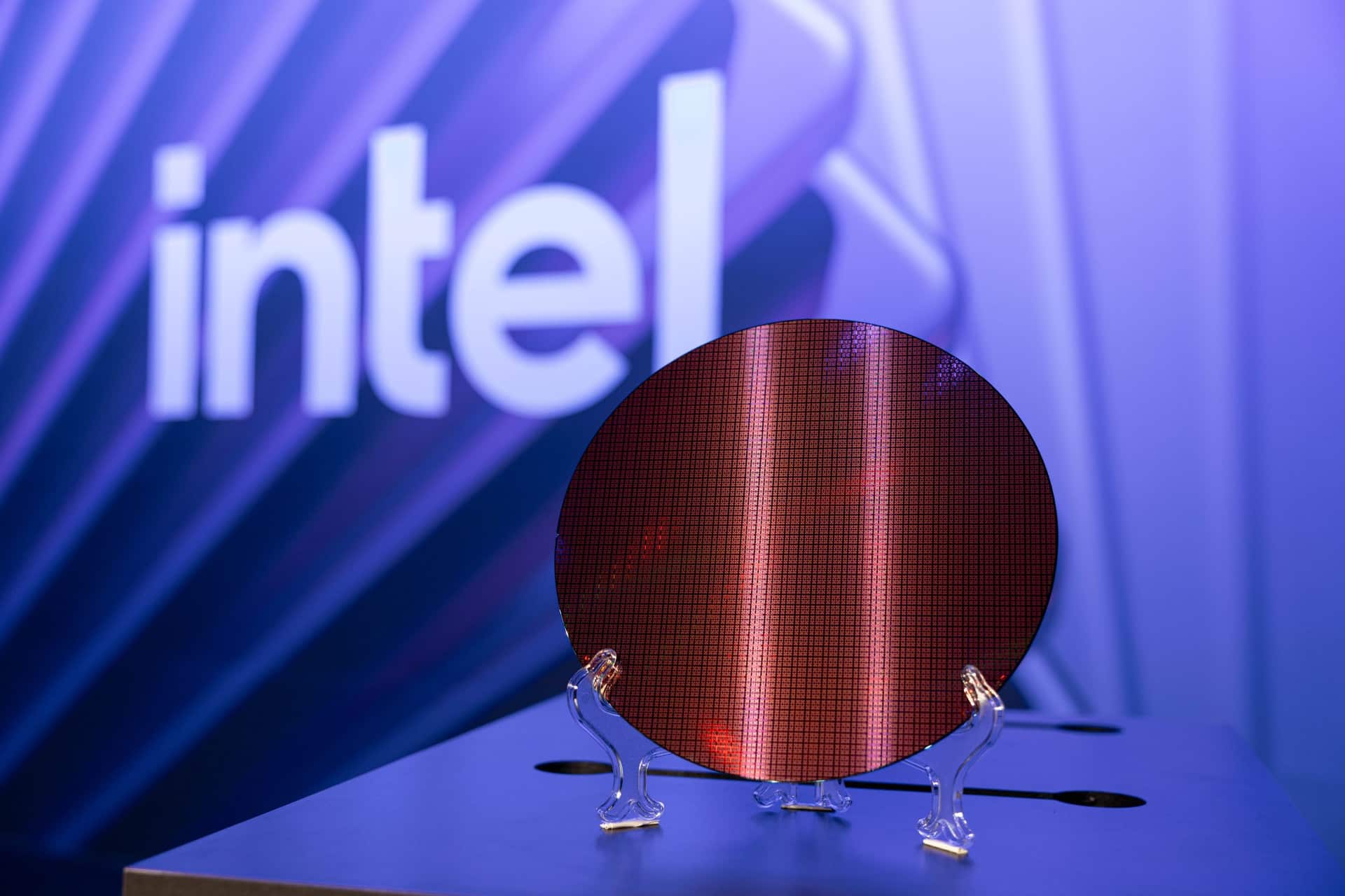 A silicon wafer is displayed on a stand in front of a blurred Intel logo and a blue, futuristic background, highlighting advancements like Intel 18A-P technology.