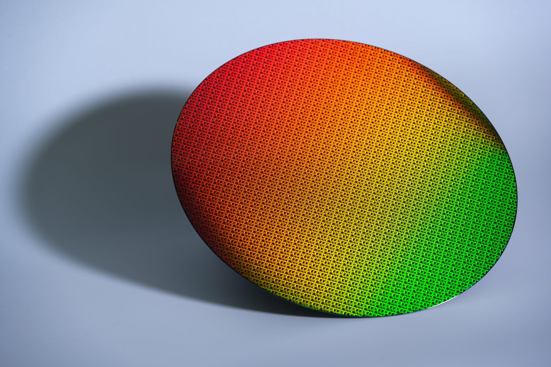 A circular silicon wafer, reminiscent of those used for Apple chips, with a grid pattern displays a gradient of colors from red and orange on one side to green on the other, casting a shadow on a light blue-gray surface.