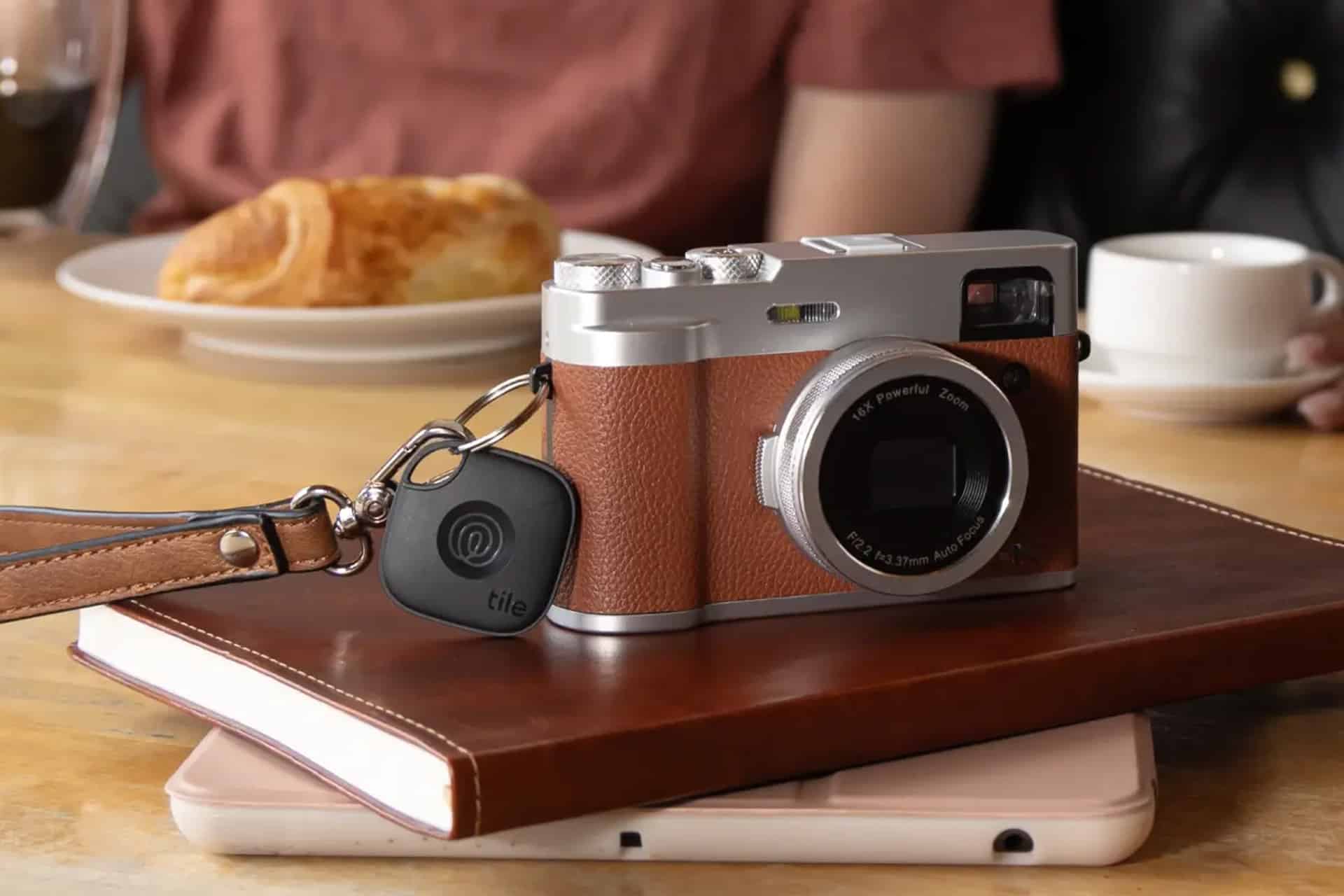 A vintage-style camera with a brown leather case and a Tile tracker keychain—perfect for Find My or Life360 location sharing—rests on a closed notebook and tablet on a table; blurred plates of food and a person are in the background.