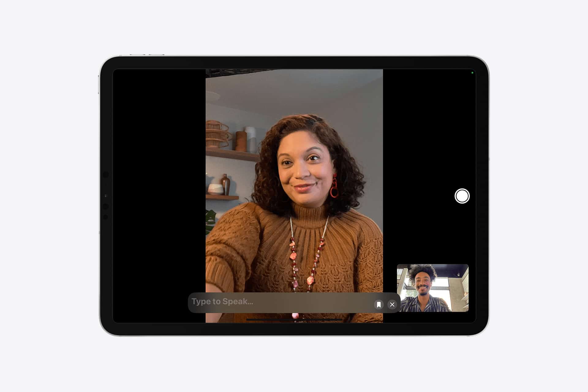 A tablet screen shows a video call between a woman in a brown sweater and a man, using Apple accessibility features like Personal Voice. The woman smiles indoors with shelves behind her; the man’s video appears in a small window at the bottom right.