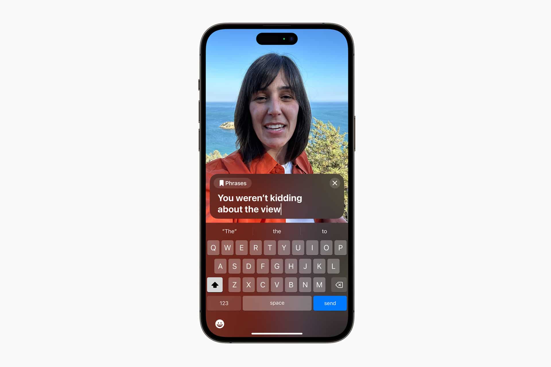 A smartphone screen shows a video call with a smiling woman outdoors, with the sea and rocks in the background. Using Apple accessibility features like Personal Voice, a message is being typed: "You weren’t kidding about the view.