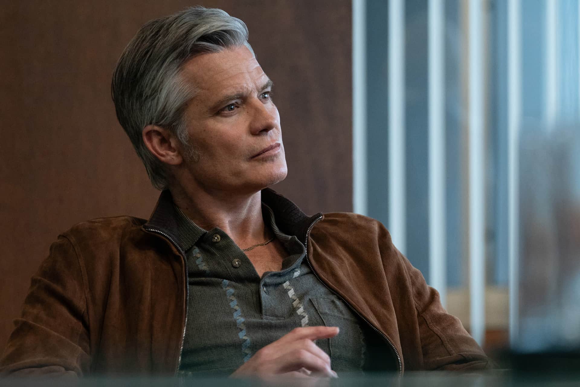 A man with gray hair sits and looks to the side, wearing a brown suede jacket over a patterned shirt. He appears thoughtful and relaxed, reminiscent of a scene from the Lucky trailer, with a blurred background of vertical lines and brown tones.