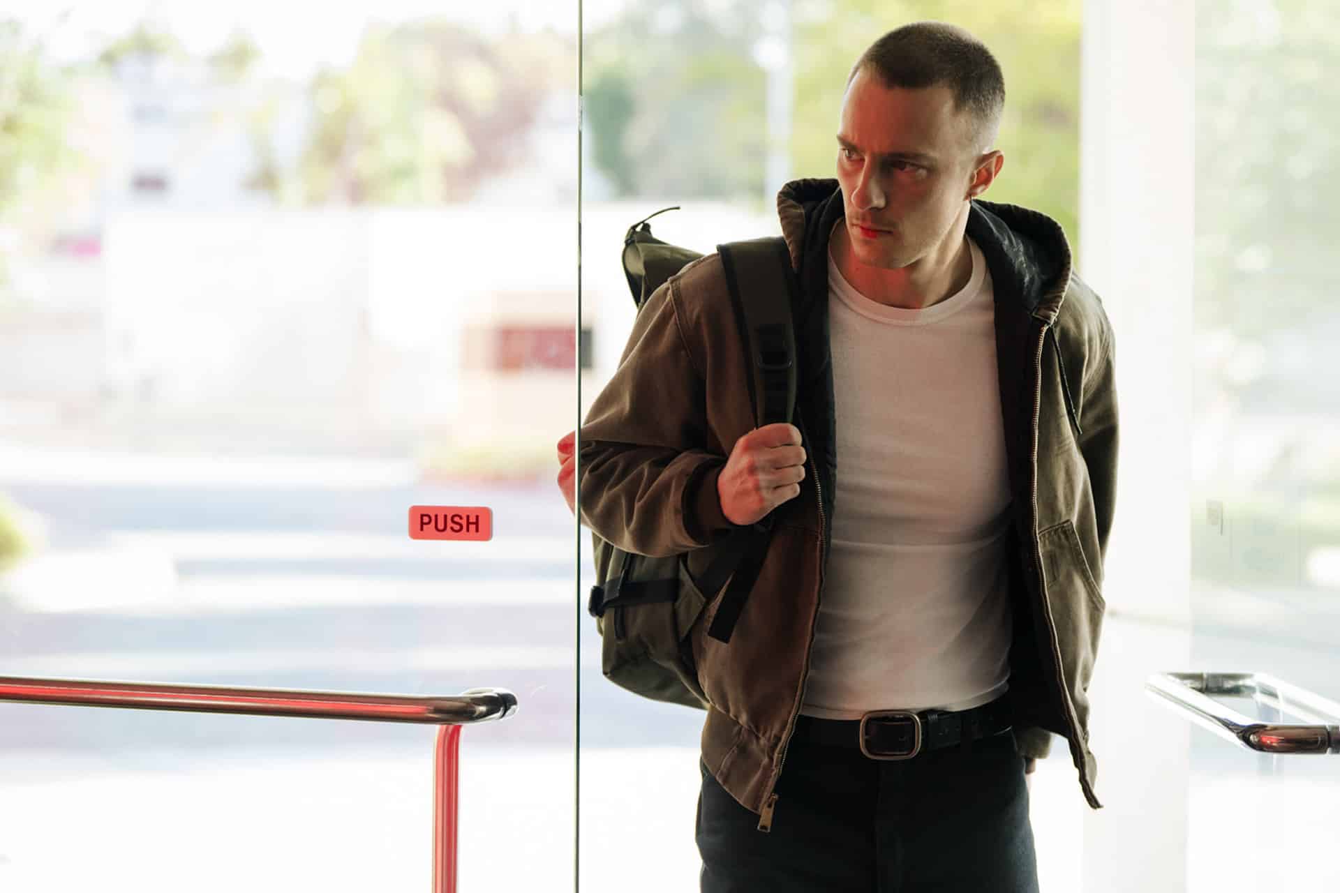 A man with a short haircut wearing a brown jacket, white t-shirt, and backpack is entering a building through a glass door marked "PUSH," looking to his right with a serious expression, as if searching for something—or someone—lucky from Apple Studios.
