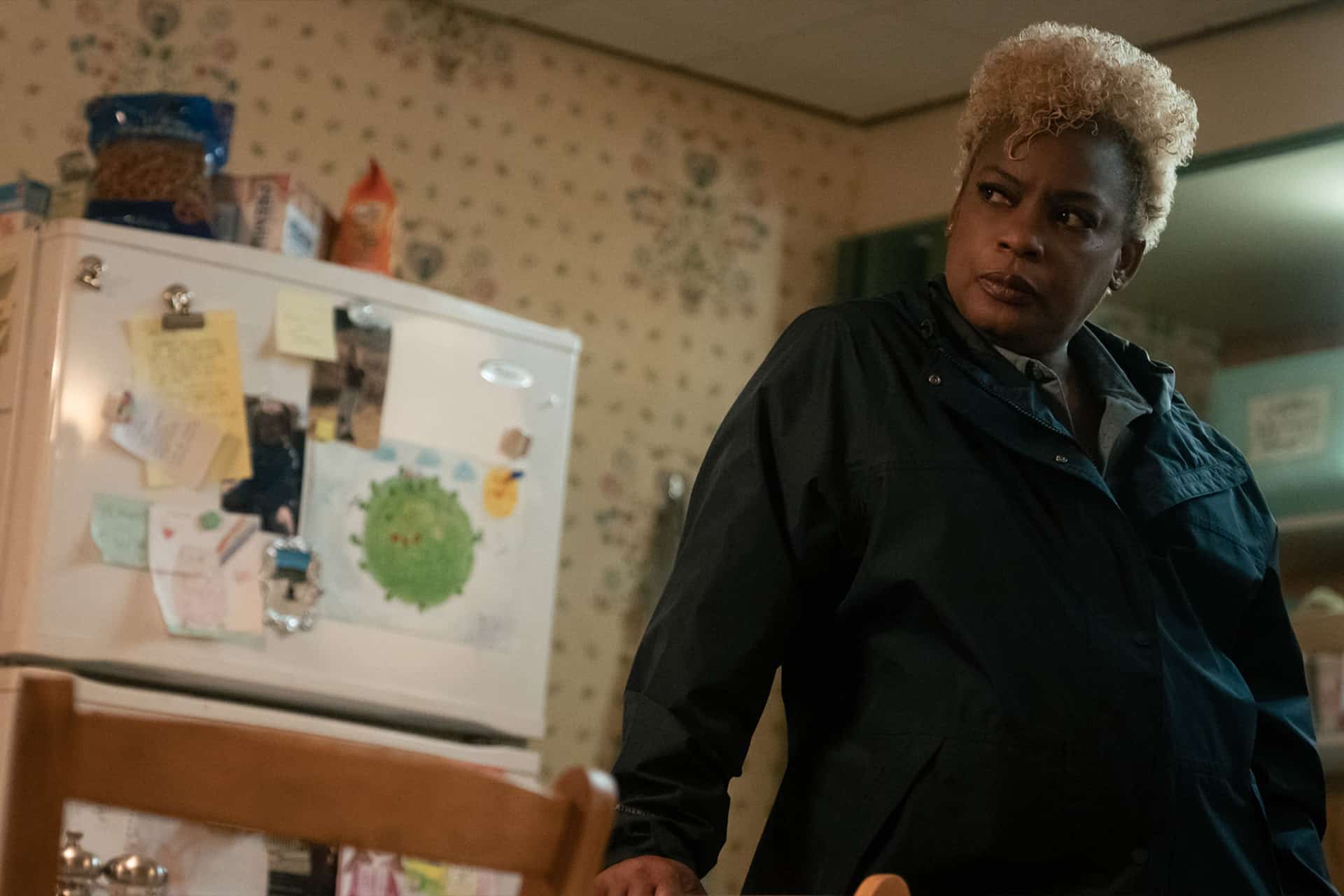 A woman with short curly blonde hair stands in a kitchen, leaning against a table and looking to the side. The refrigerator behind her is covered with magnets, papers, and children's artwork—much like a scene from the Lucky trailer.