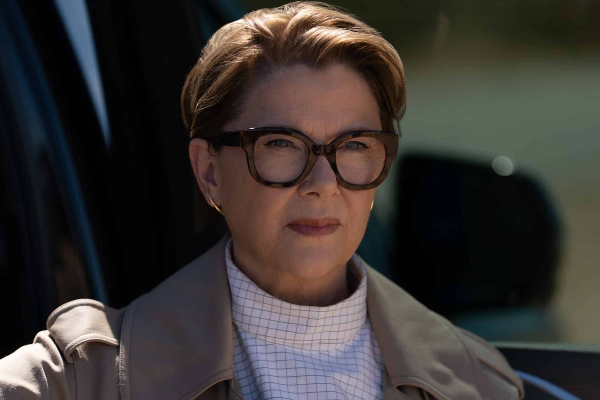 A woman with short light brown hair, large dark glasses, and a beige trench coat over a checkered shirt looks ahead seriously. She is outdoors, possibly beside a vehicle in a scene from the Lucky trailer by Apple TV and Apple Studios.