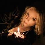 A woman with blonde hair and a serious expression lies in dim light, holding a lit lighter that illuminates her face and part of her black clothing, reminiscent of Anya Taylor-Joy in an Apple Studios' Lucky trailer.