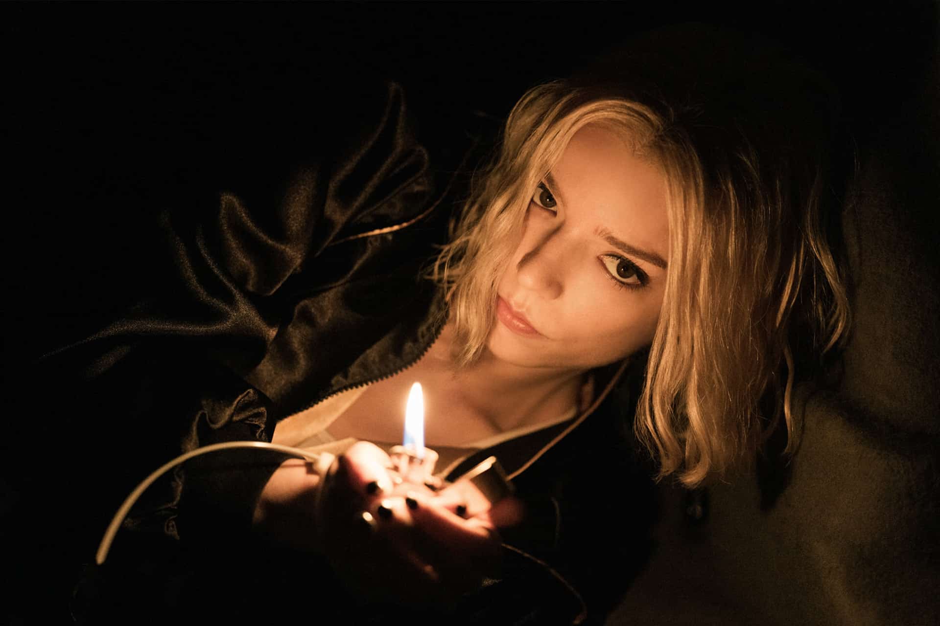 A woman with blonde hair and a serious expression lies in dim light, holding a lit lighter that illuminates her face and part of her black clothing, reminiscent of Anya Taylor-Joy in an Apple Studios' Lucky trailer.