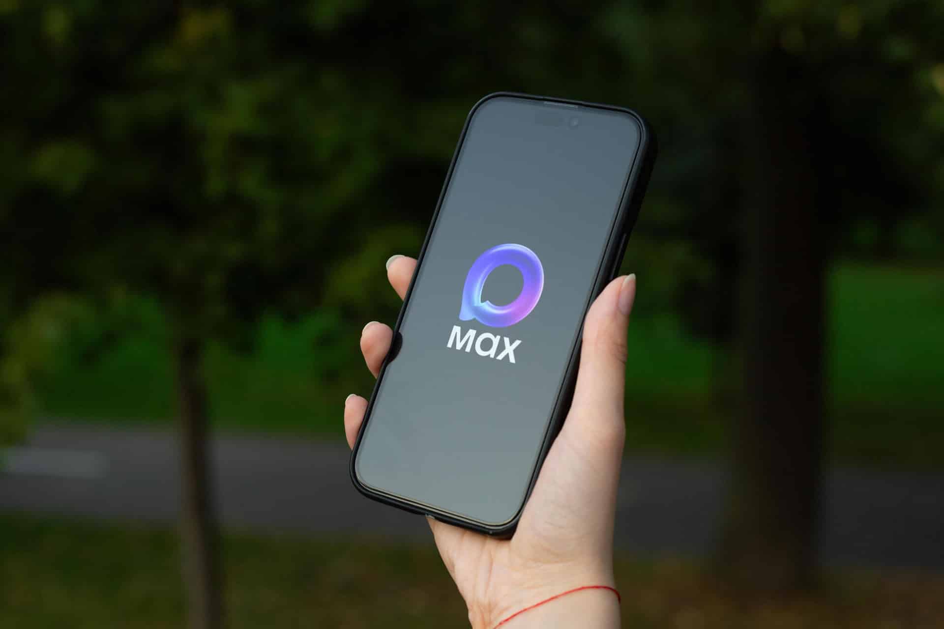 A hand holds a smartphone outdoors, displaying a dark screen with the "Max" streaming service logo in the center. The background is blurred with green trees and a pathway, following recent headlines like "Apple removes Max" from its Apple Russia store.