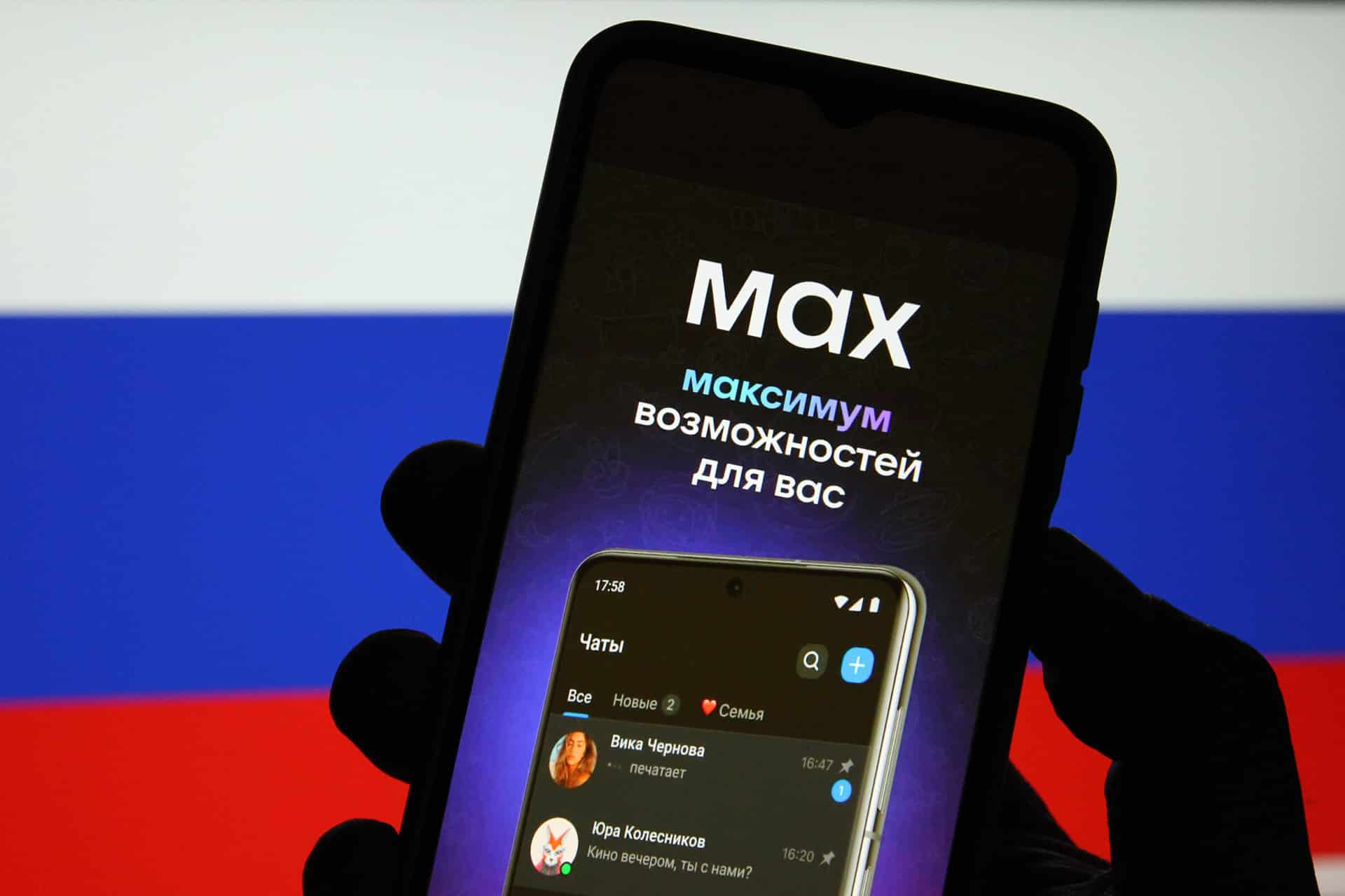 A hand holds a smartphone displaying a Russian messaging app with Cyrillic text, against a blurred background of the Russian flag. The screen shows chats and the words “максимум возможностей для вас” (“maximum possibilities for you”).