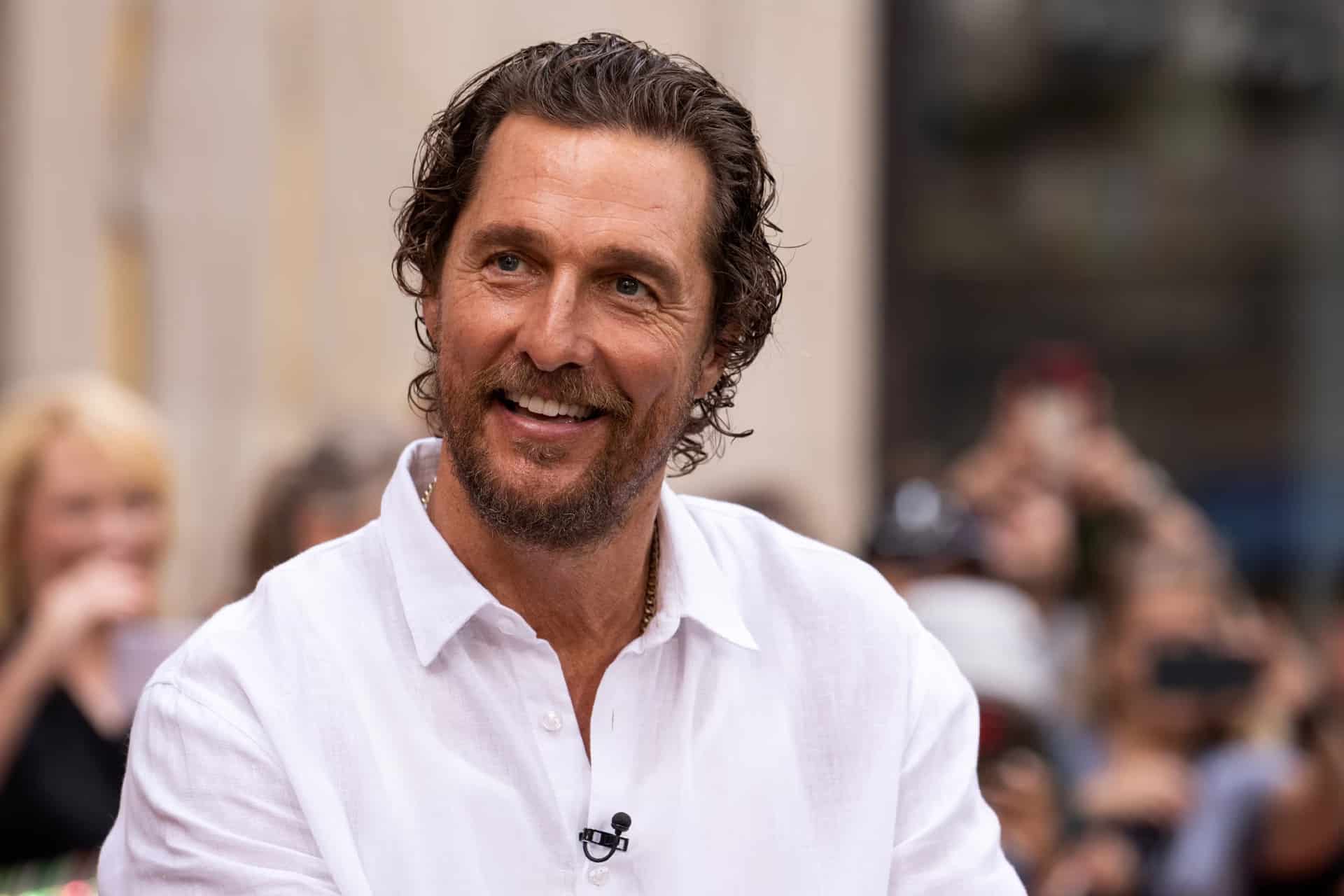 A man with wavy brown hair and a beard, wearing a white shirt, smiles while sitting outdoors, resembling Matthew McConaughey in an Apple TV scene, with a blurred crowd and buildings in the background.