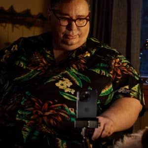 A man wearing glasses and a black floral shirt sits indoors, looking at a smartphone on a stand in front of him, possibly using Apple accessibility features like Personal Voice. Soft, warm lighting and an evening-blue-lit window set the background.