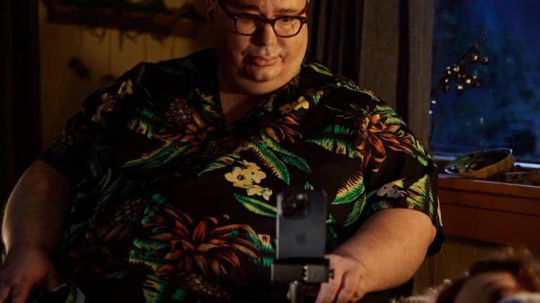 A man wearing glasses and a black floral shirt sits indoors, looking at a smartphone on a stand in front of him, possibly using Apple accessibility features like Personal Voice. Soft, warm lighting and an evening-blue-lit window set the background.