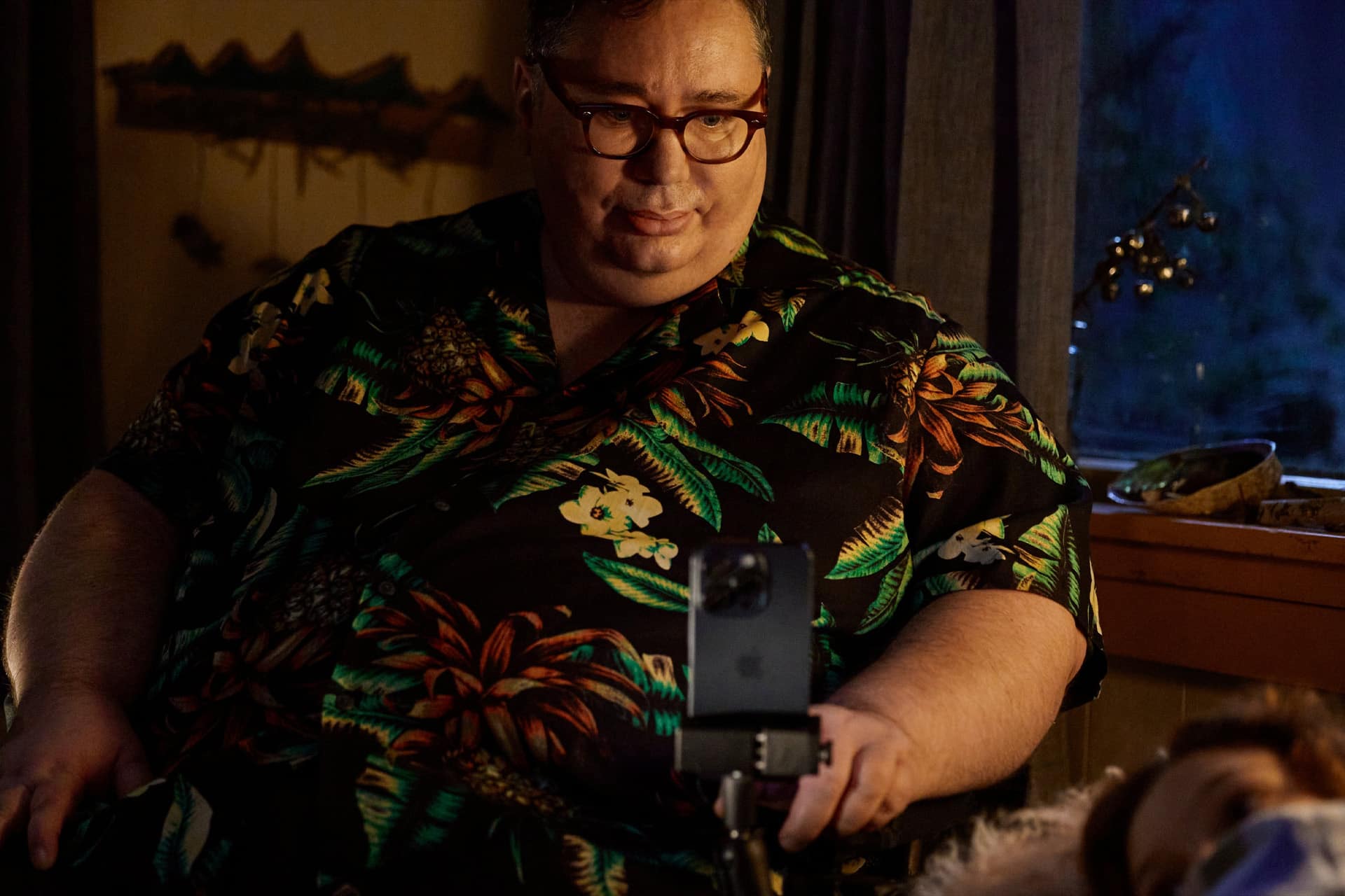 A man wearing glasses and a black floral shirt sits indoors, looking at a smartphone on a stand in front of him, possibly using Apple accessibility features like Personal Voice. Soft, warm lighting and an evening-blue-lit window set the background.