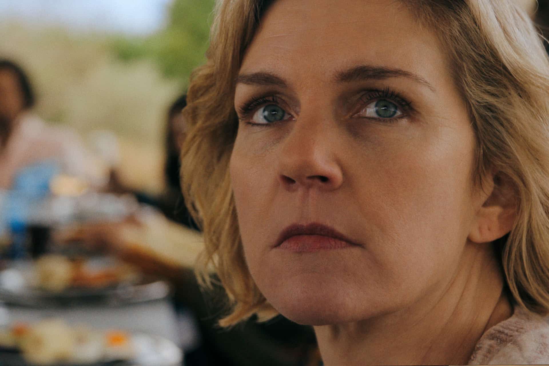 A woman with short blonde hair looks slightly upward with a serious expression. Behind her, a blurred background suggests a lively scene at the Gotham Television Awards, with people and plates of food on a table.