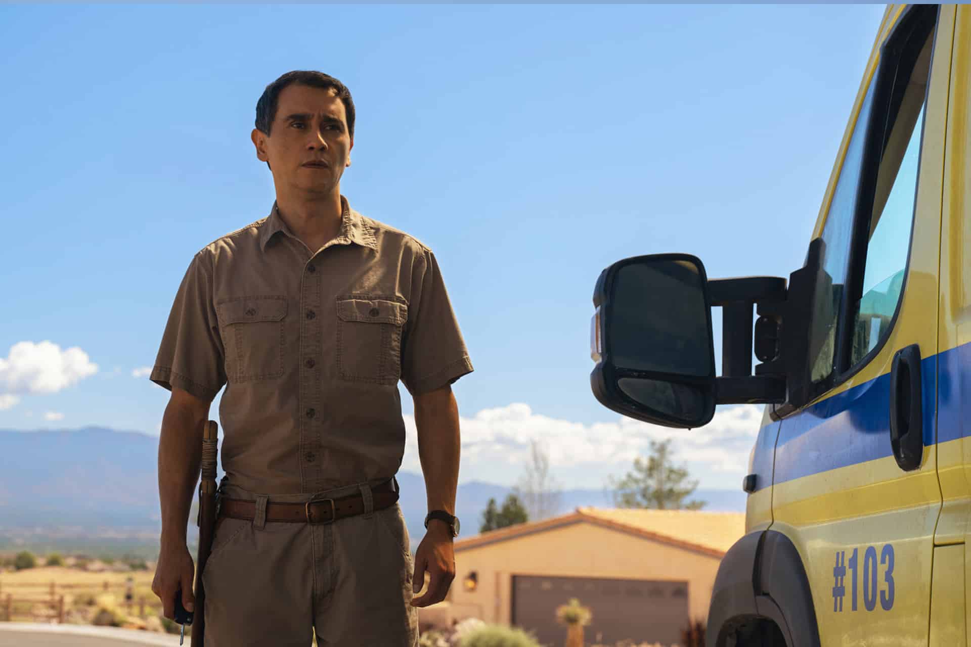 A man in a tan uniform stands outdoors holding a shotgun beside a yellow emergency vehicle with "#103" on the side. Desert scenery, mountains, and houses are visible under a blue sky—like a scene from acclaimed Apple Originals.
