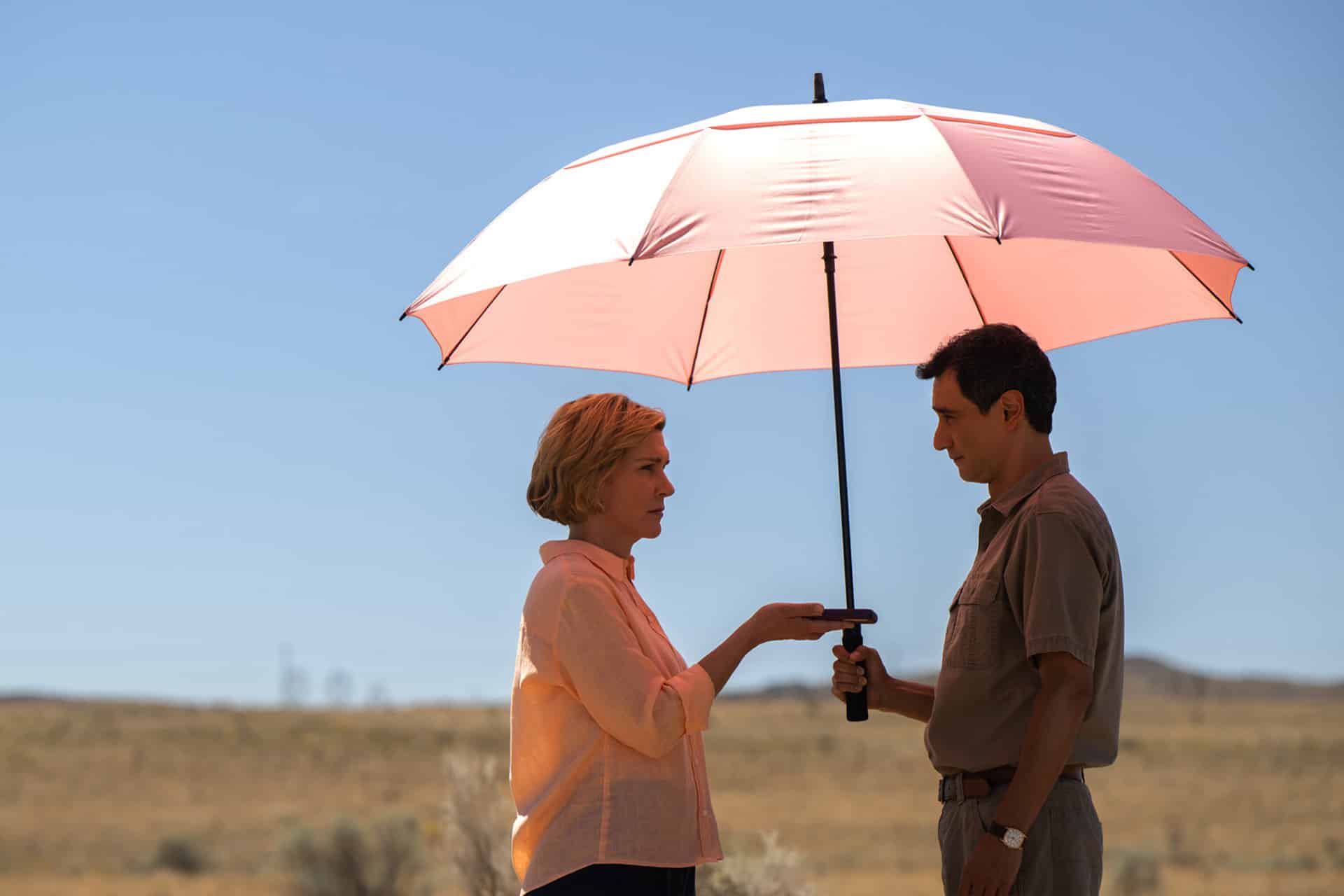 A woman and a man stand facing each other outdoors under a large pink umbrella—Apple Originals style—with the woman holding the handle and the man steadying the pole, set against a clear blue sky worthy of a Gotham Television Awards scene.