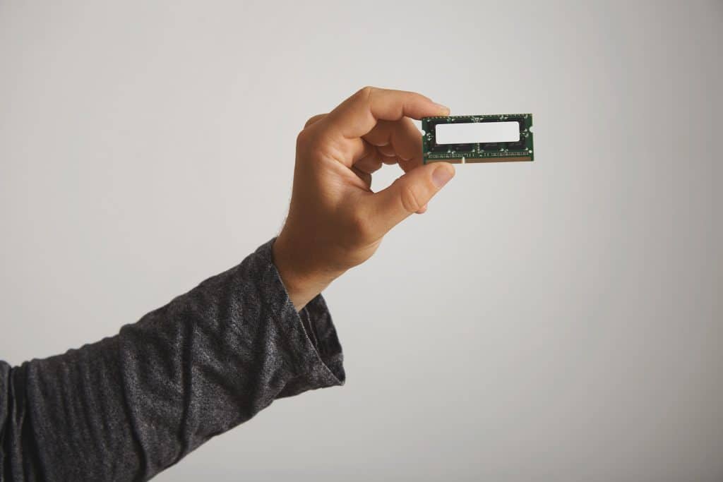 A person wearing a long-sleeve dark shirt holds a DRAM memory module between their thumb and fingers, showcasing its memory chips against a plain light background.