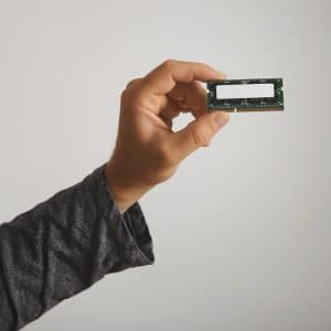 A person wearing a long-sleeve dark shirt holds a DRAM memory module between their thumb and fingers, showcasing its memory chips against a plain light background.