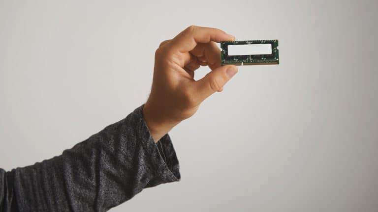 A person wearing a long-sleeve dark shirt holds a DRAM memory module between their thumb and fingers, showcasing its memory chips against a plain light background.
