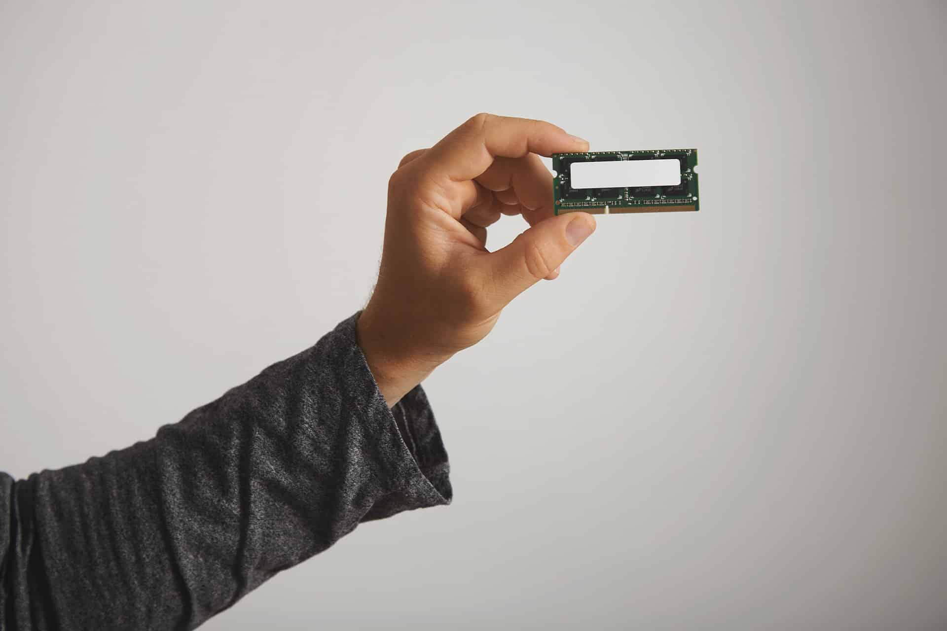 A person wearing a long-sleeve dark shirt holds a DRAM memory module between their thumb and fingers, showcasing its memory chips against a plain light background.