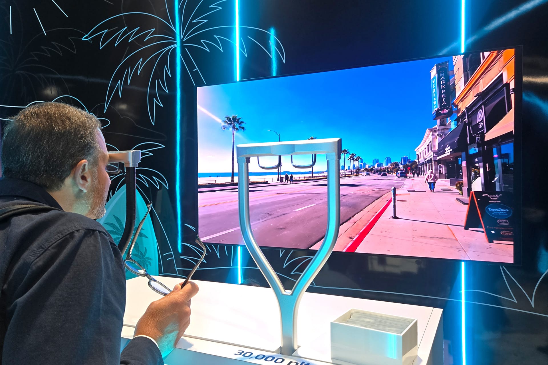 A man closely examines a large, high-definition Samsung XR display showing a bright, sunny street scene at a technology exhibit, with modern display equipment in the foreground and neon palm tree outlines on the walls.