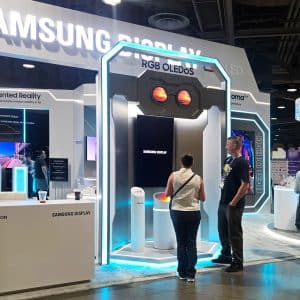 People explore Samsung Display exhibits at a tech convention. Bright lights illuminate screens and Samsung XR displays, with visitors interacting and viewing information at various booths. The atmosphere is modern and high-tech.