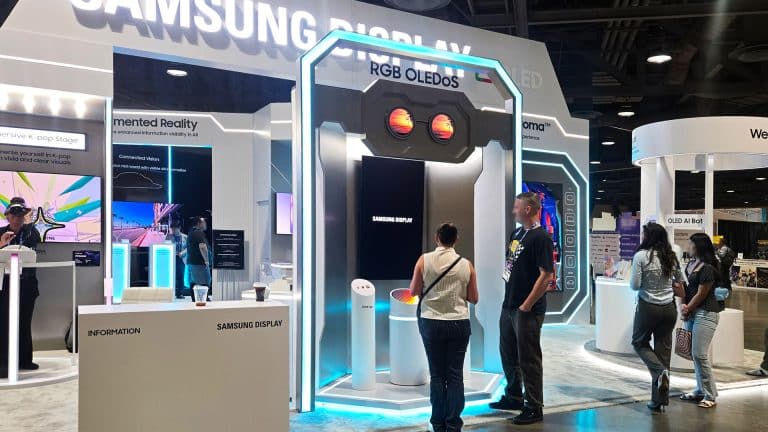 People explore Samsung Display exhibits at a tech convention. Bright lights illuminate screens and Samsung XR displays, with visitors interacting and viewing information at various booths. The atmosphere is modern and high-tech.